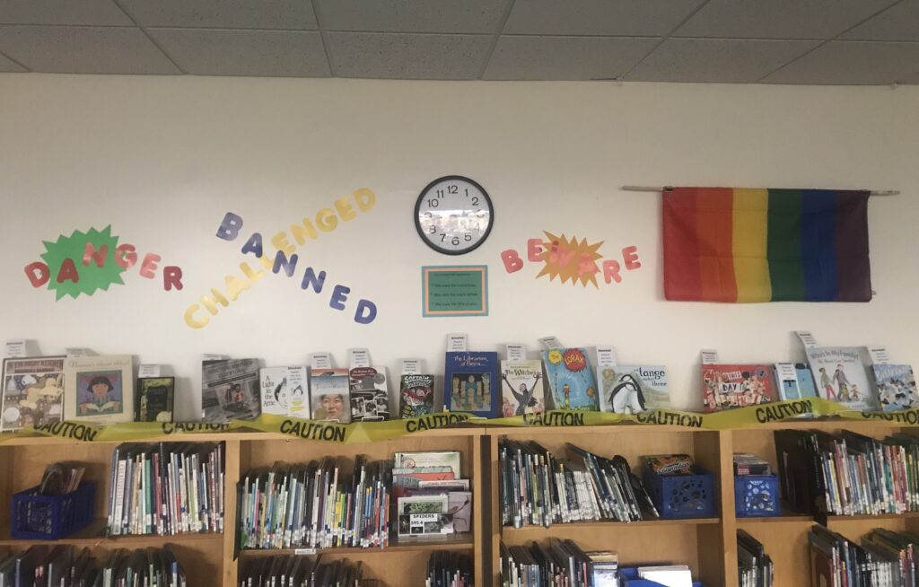 DANGER! BEWARE! CAUTION! Challenged and Banned Books on display below a Pride flag. Including titles like "And Tango Makes Three," "A Light in the Attic," "In the Night Kitchen," "The Watson's Go to Montgomery," "The Lorax," and "Nasreen's Secret School." 